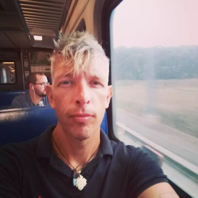 The image shows a man with a distinctive hairstyle, possibly on a train. He's taking a selfie, looking directly at the camera. In the background, another person is visible, seemingly also on the train. The overall impression is casual, suggesting a moment captured during a journey.